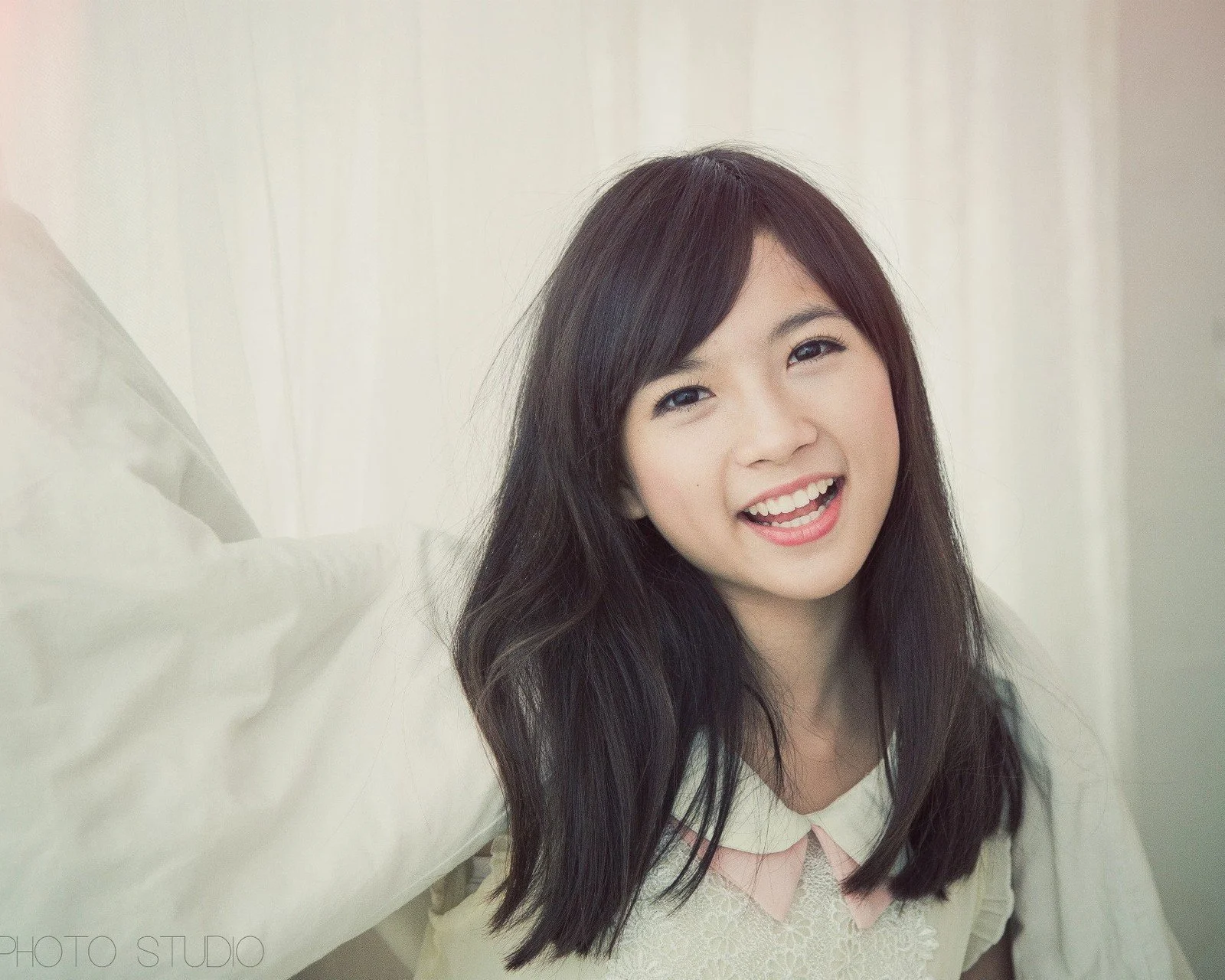 긴 머리의 여성이 흰색 상의를 입고 밝은 커튼과 베개 앞에서 미소 짓고 있습니다.