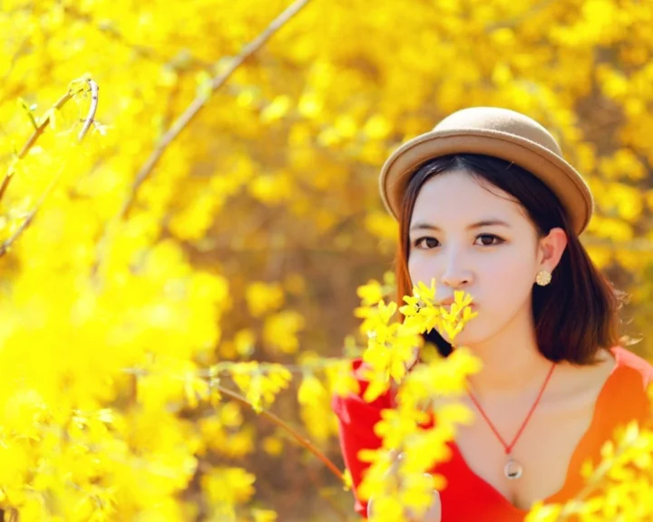 帽子をかぶった女性が、赤い服を着て黄金色の秋の葉に囲まれて立っています。