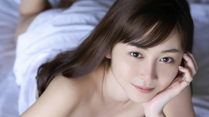 一位棕髮成年女性托腮面向鏡頭微笑，背景是柔和的床鋪。