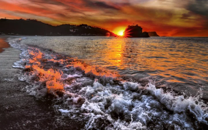 夕阳映照着海滩与翻涌的海浪，远处是 silhouetted 岬角与绚丽晚霞。