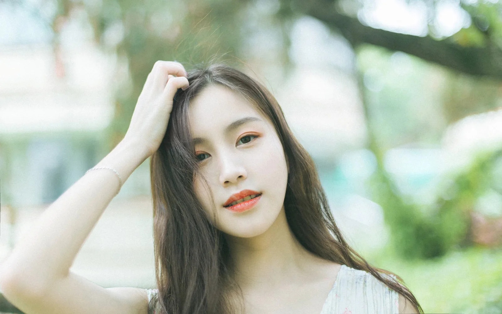 一名长发女性在户外绿意背景前轻抚头发并看向镜头。