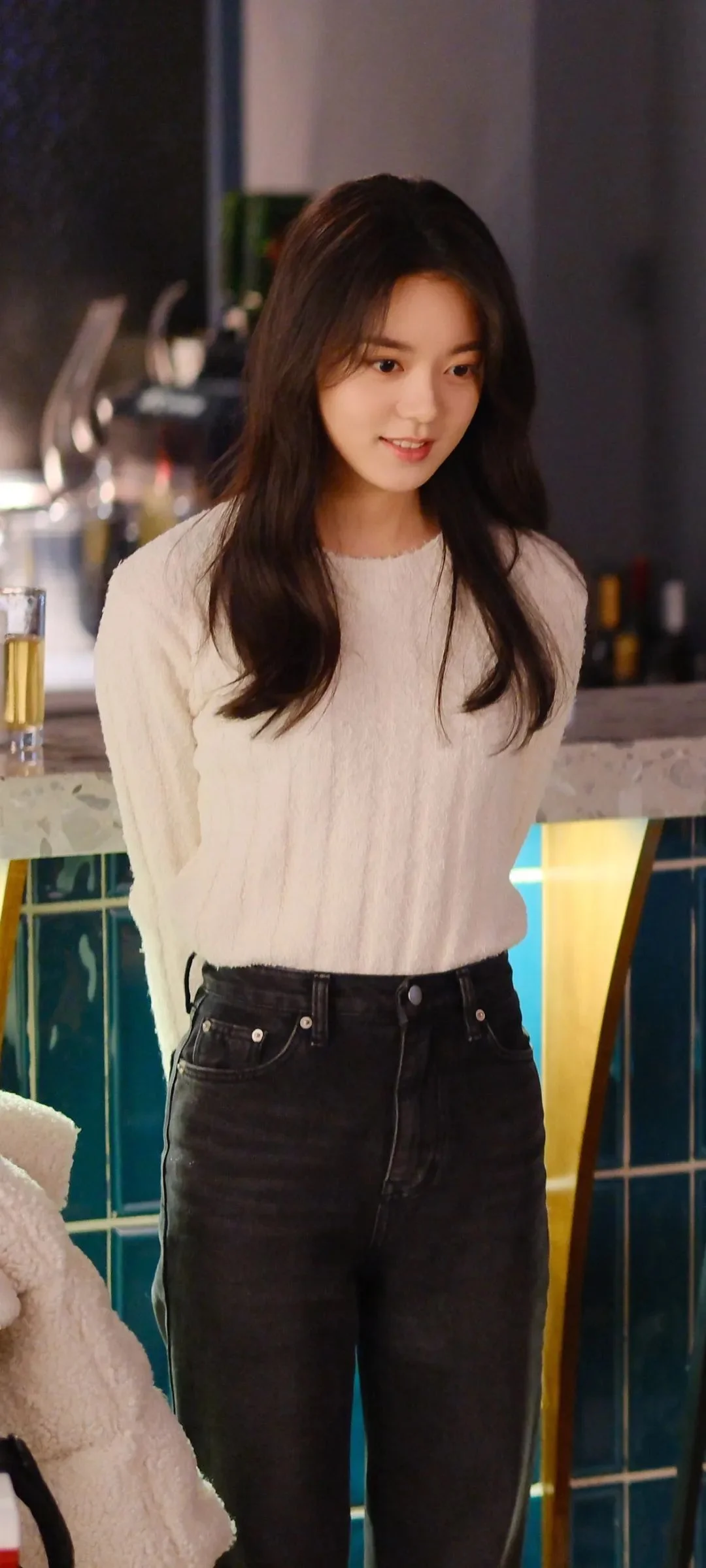 긴 머리의 여성이 밝은 색 니트를 입고 실내에 서 있습니다.