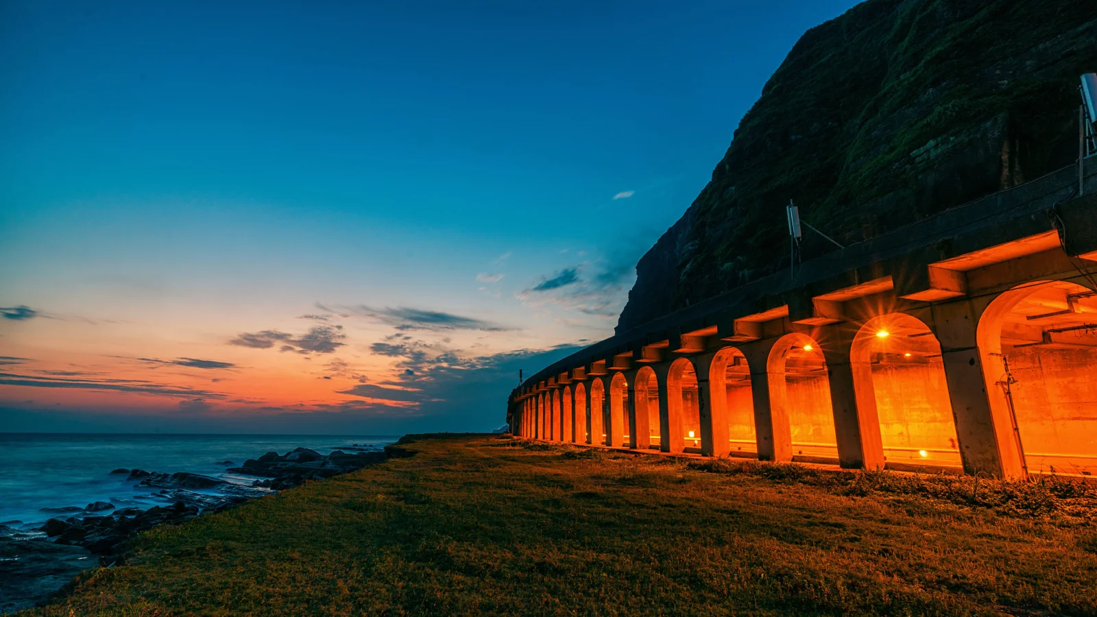 夕日に照らされた海岸と山肌に沿うオレンジ色の照明のトンネルが印象的な風景です。
