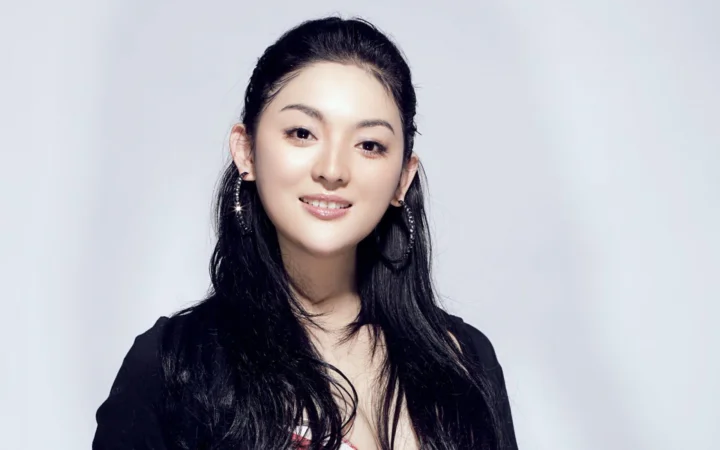 一位长发女性身穿黑色上衣，在浅灰色背景前微笑面对镜头。