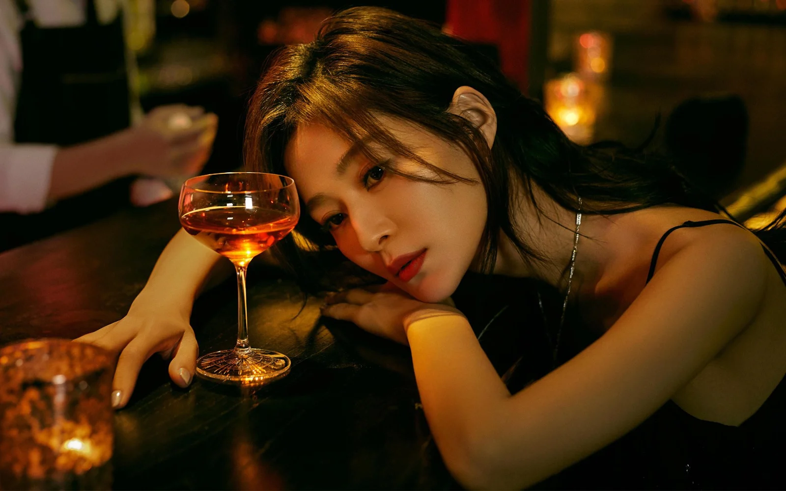 一名女子靠在桌边，身旁放着一杯葡萄酒，背景光线昏暗。