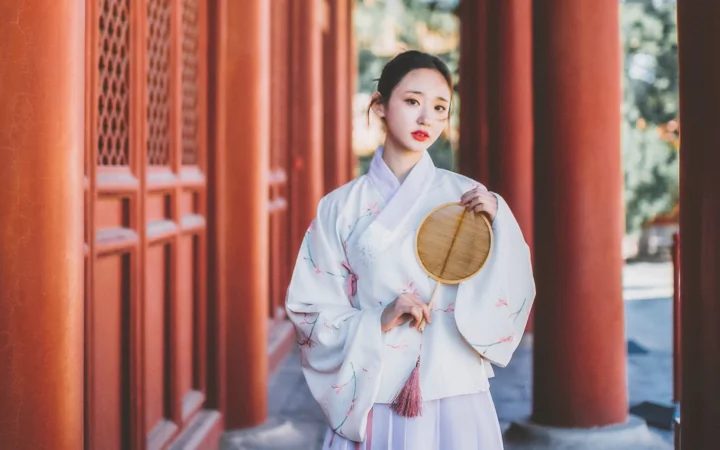 一名身穿浅色传统服饰、手持圆扇的女子站在红色柱廊之间。