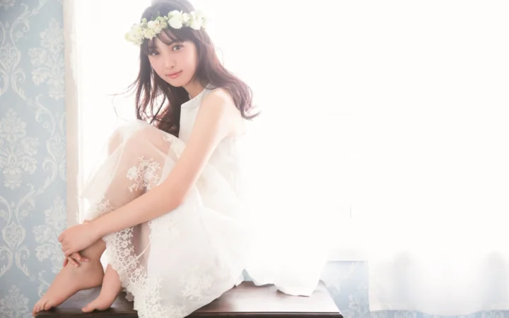一名戴着花环、身穿白裙的女性坐在明亮窗边。