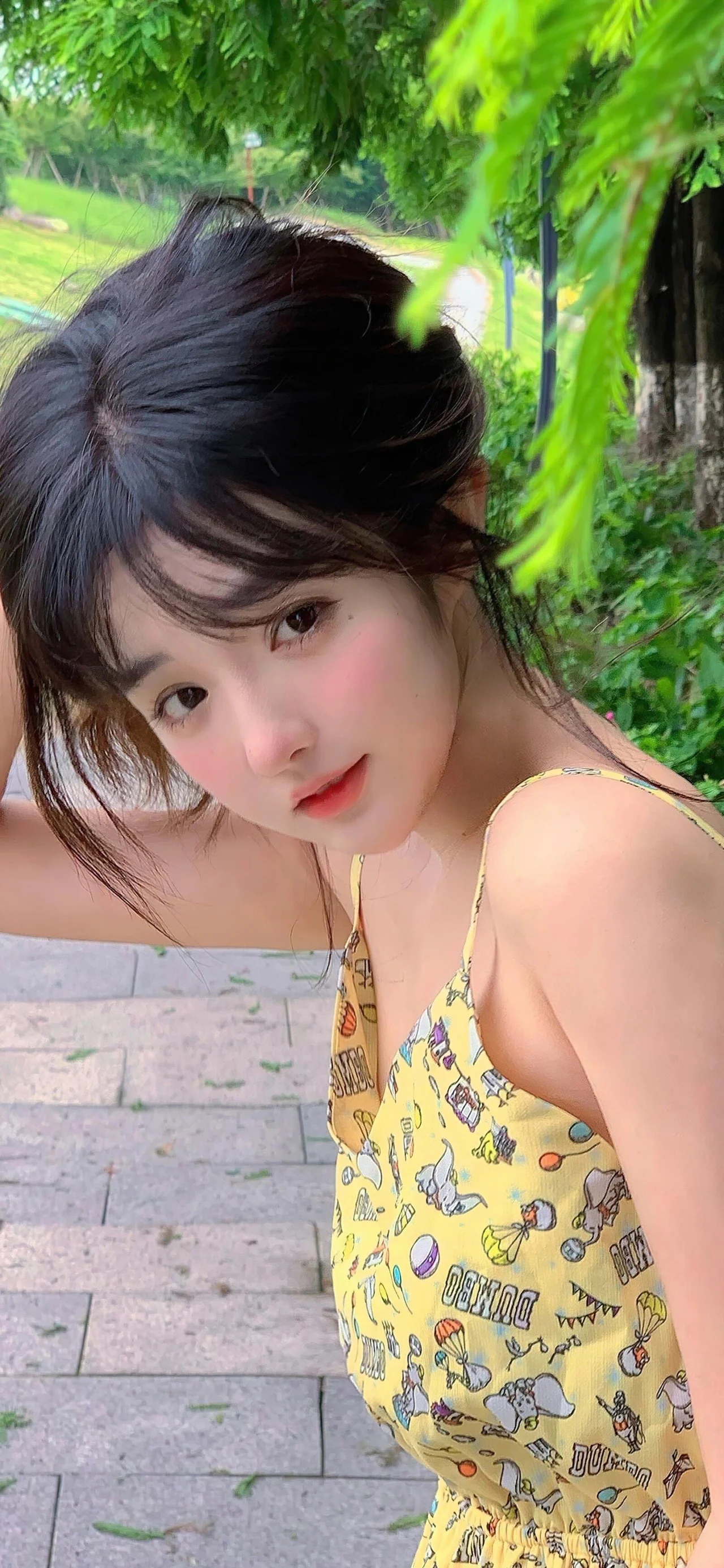 一名黑发女子穿着黄色图案连衣裙，在绿叶环绕的户外步道旁看向镜头。