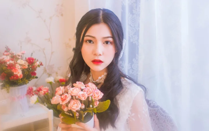 一名成年女性在室内手捧粉色花束，身后可见花卉与浅色窗帘。