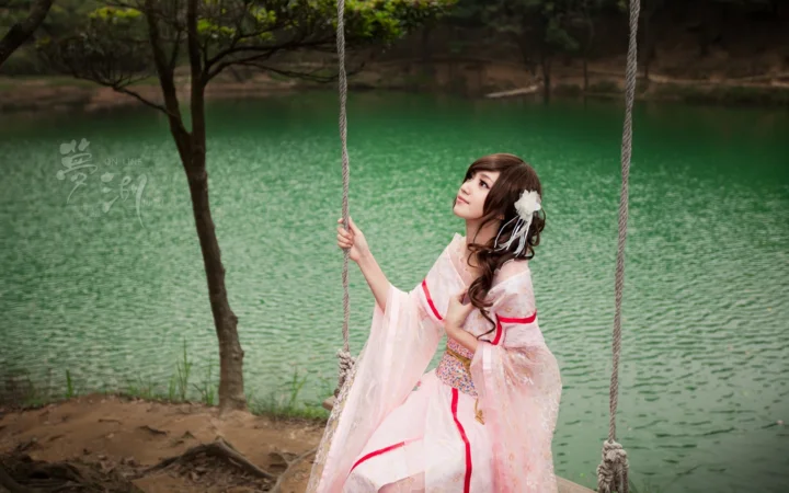一名身穿粉色传统服饰的女子坐在湖畔秋千上，背景是绿色湖水和树木。
