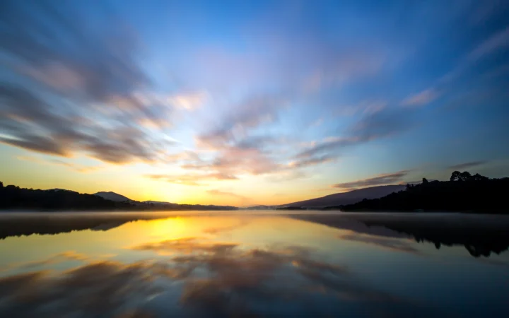 夕阳映照在平静湖面上，远处山影与天空中的云彩共同构成宁静的自然景色。