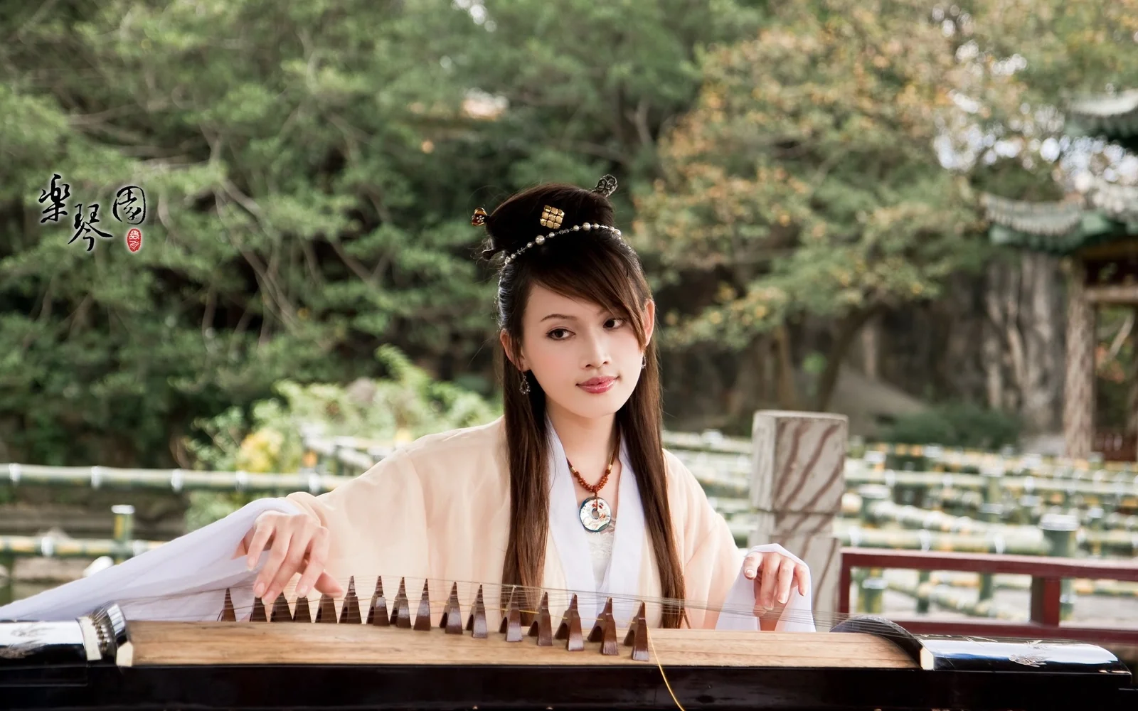 伝統衣装を着た成人女性が、木々に囲まれた庭園で古筝を演奏しています。