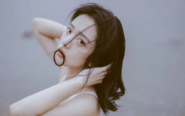 一名长发女性在冷灰色背景前侧头看向镜头，双手置于头颈附近。