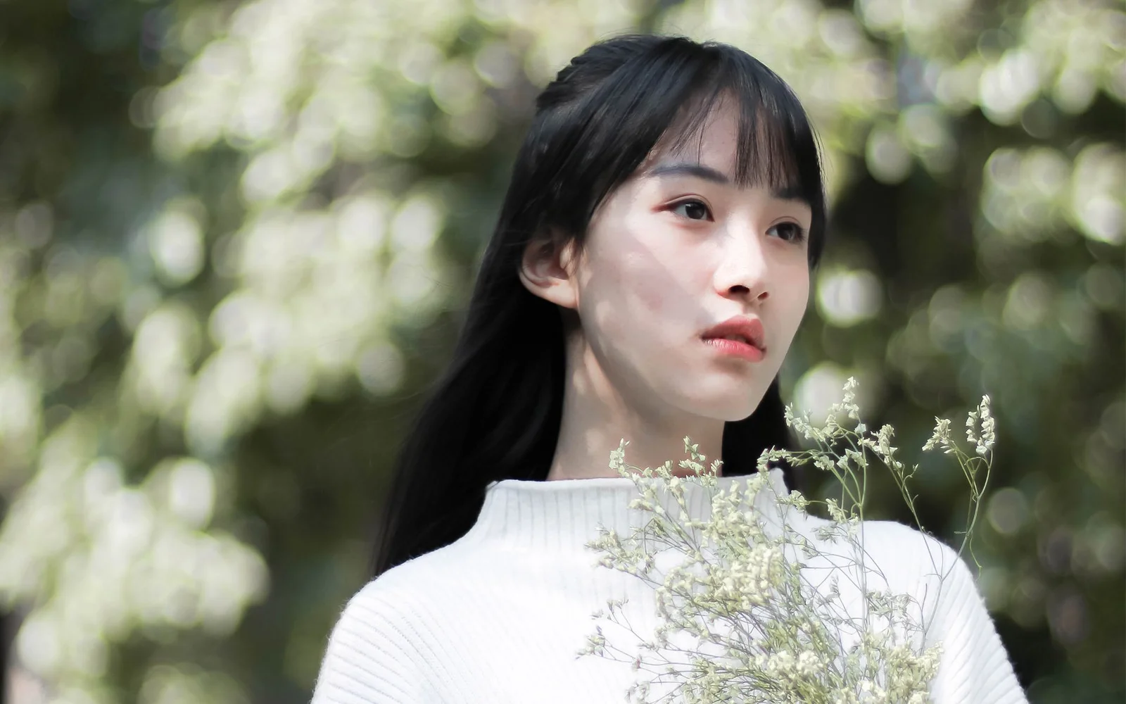 一名黑发女子身穿白色上衣站在林间，身前映入几枝细小的白色野花。