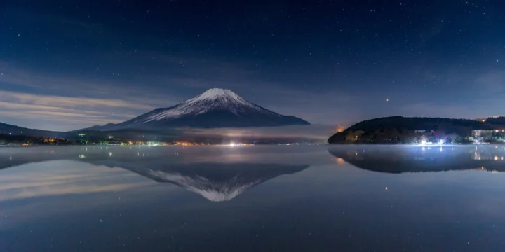夜空下的雪山倒映在平静湖面上，岸边点缀着微弱灯光。