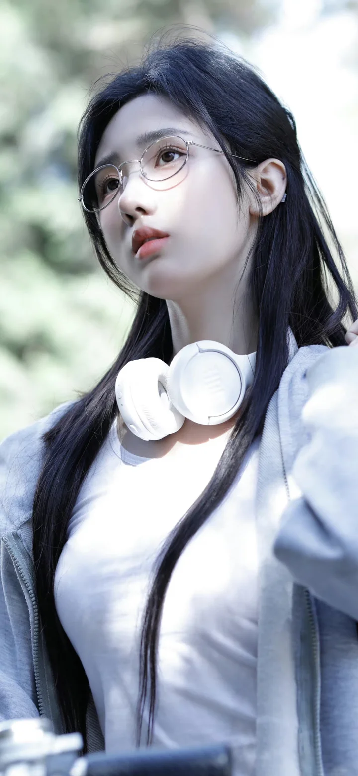 一名戴圆框眼镜的长发女性站在户外，白色耳机挂在颈部，背景为虚化的绿色植物。