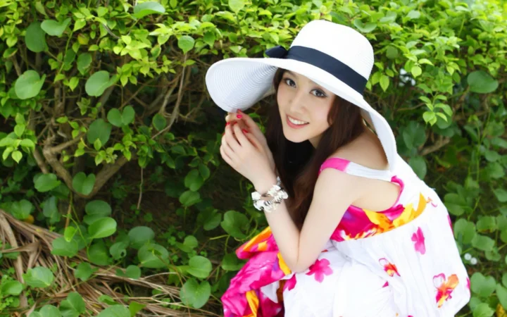 一位戴着白色宽檐帽的成年女性在绿叶背景前微笑。
