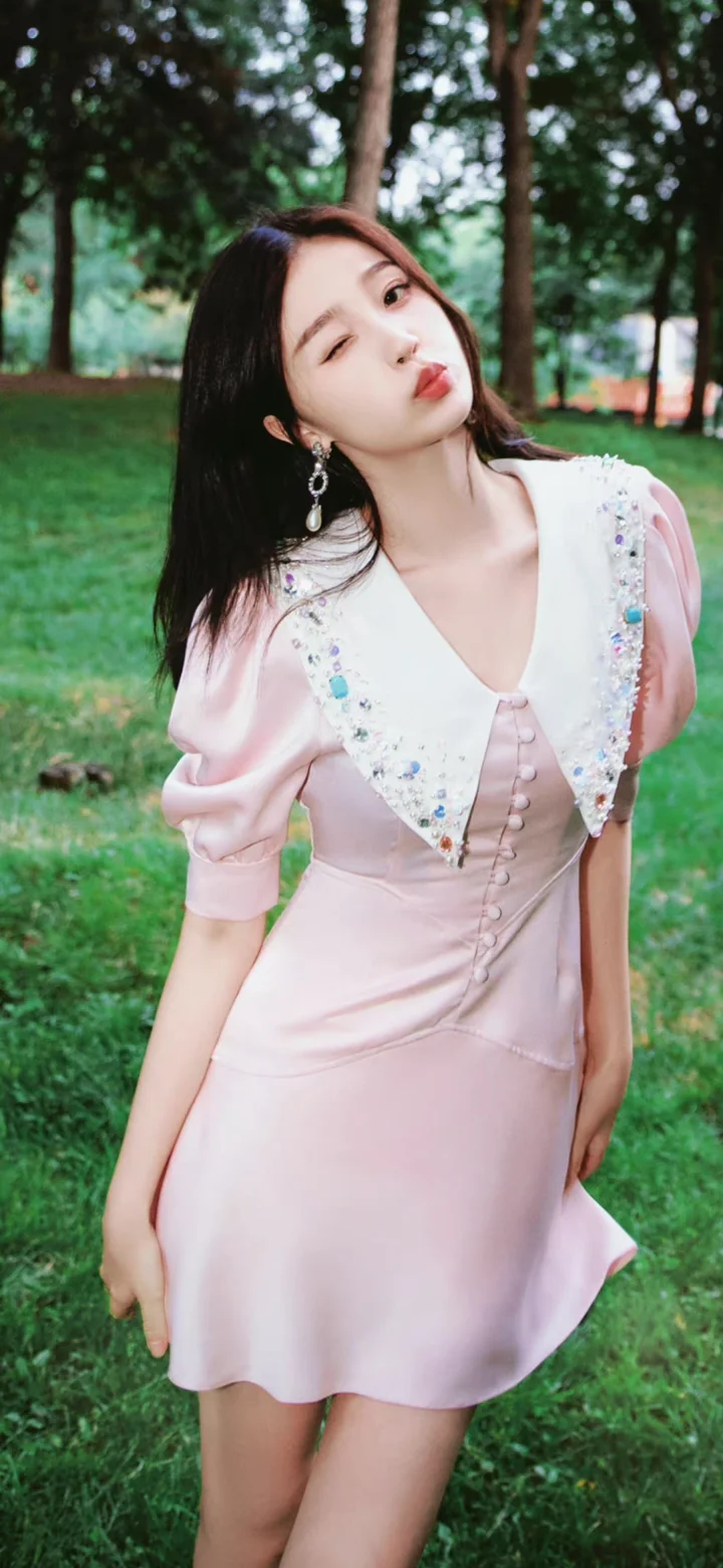 ピンクのワンピースを着た女性が、緑の木々に囲まれた屋外の芝生でカメラに向かってウインクしている。