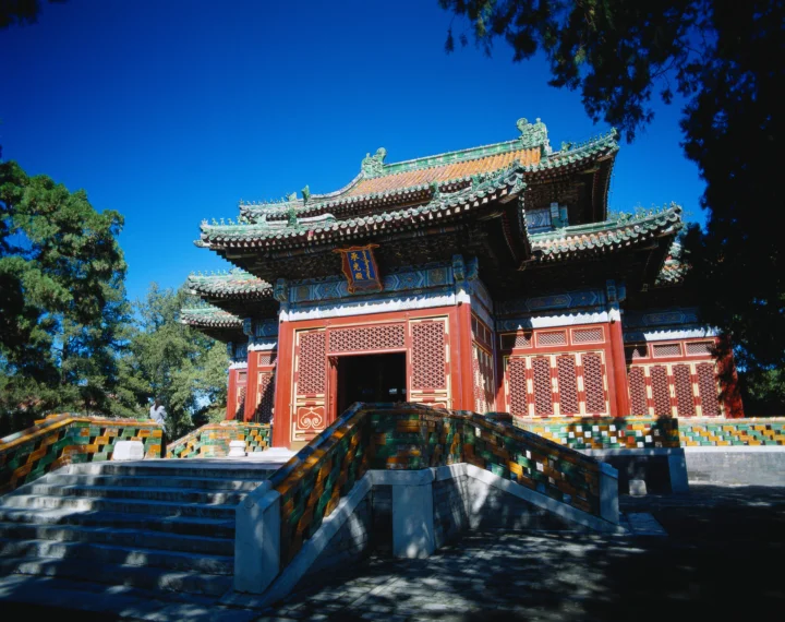 一座红色中式古建筑坐落在石阶之上，周围有树木，背景是晴朗的深蓝色天空。