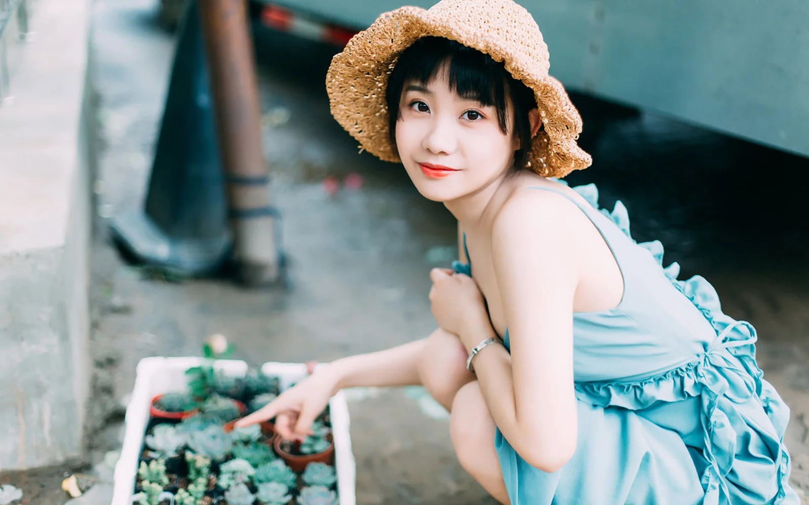 一名戴草帽、穿蓝色连衣裙的女性在户外花盆旁回头看向镜头。