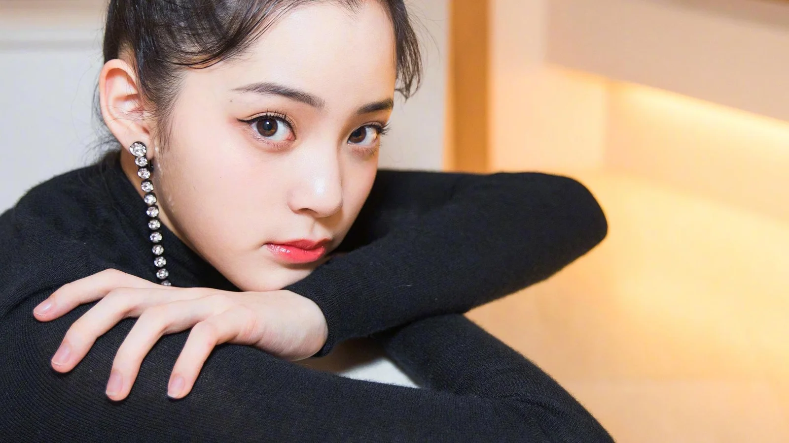黒いセーターを着た成人女性が腕に寄りかかりながらカメラを見つめ、背景には暖かな室内の光が見えます。