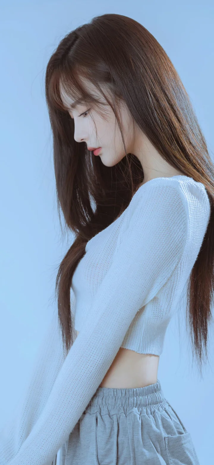 一名長髮女子側身站在淺藍色背景前，穿著白色針織上衣和灰色褲子。
