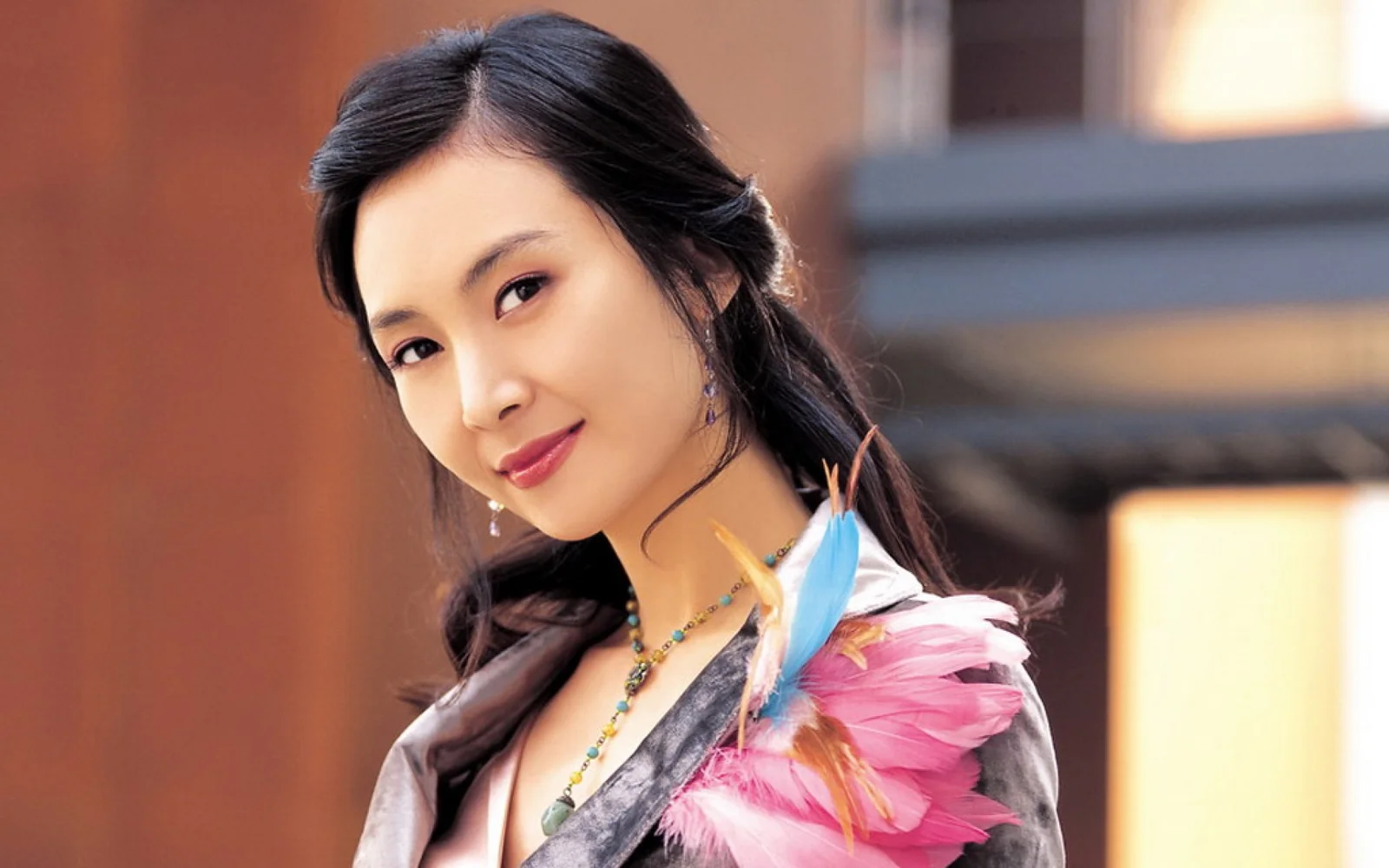 화려한 깃털 장식을 한 성인 여성이 흐릿한 건물 배경 앞에서 옆모습으로 미소 짓고 있다.