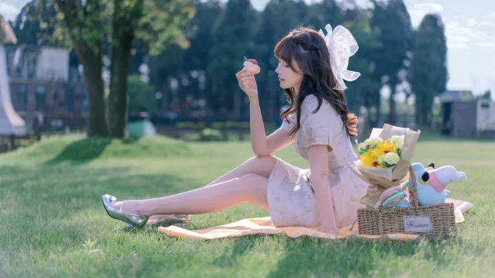 一位成年女性坐在草地上的野餐垫上，手持饮料，身旁放着花束、篮子和玩偶。