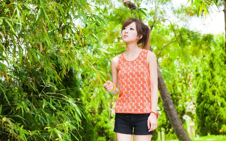 一名穿橙色背心和黑色短裤的女子站在竹林和绿树之间。