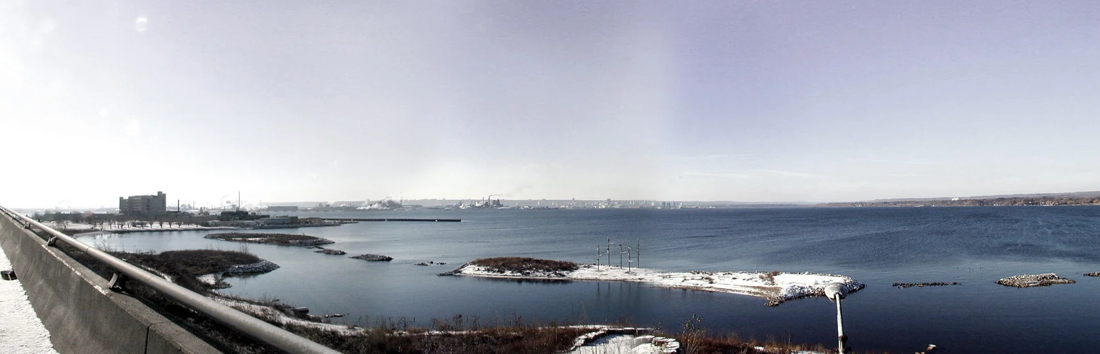冬の湾に浮かぶ雪に覆われた小島と遠くの建物、手前の橋を捉えた panorámicaです。