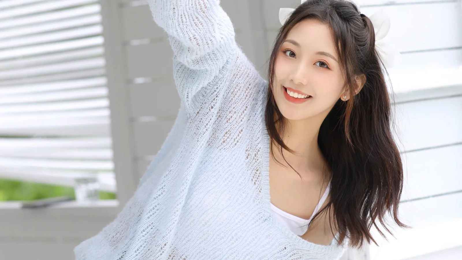 一位长发女性穿着白色针织衫，在明亮窗边微笑。