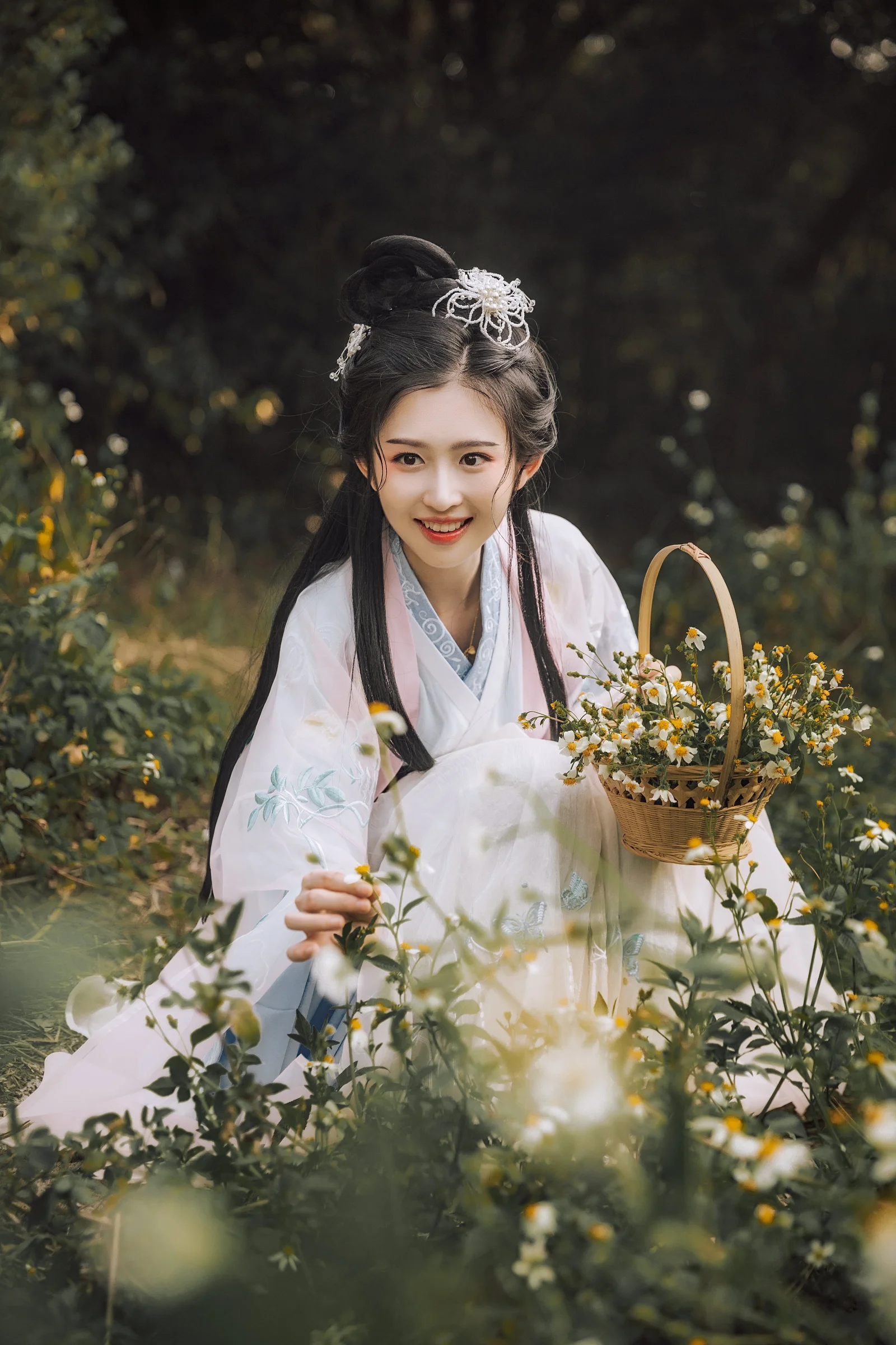 흰색 전통 의상을 입은 여성이 들꽃 사이에 앉아 꽃바구니와 나뭇가지를 들고 있습니다.