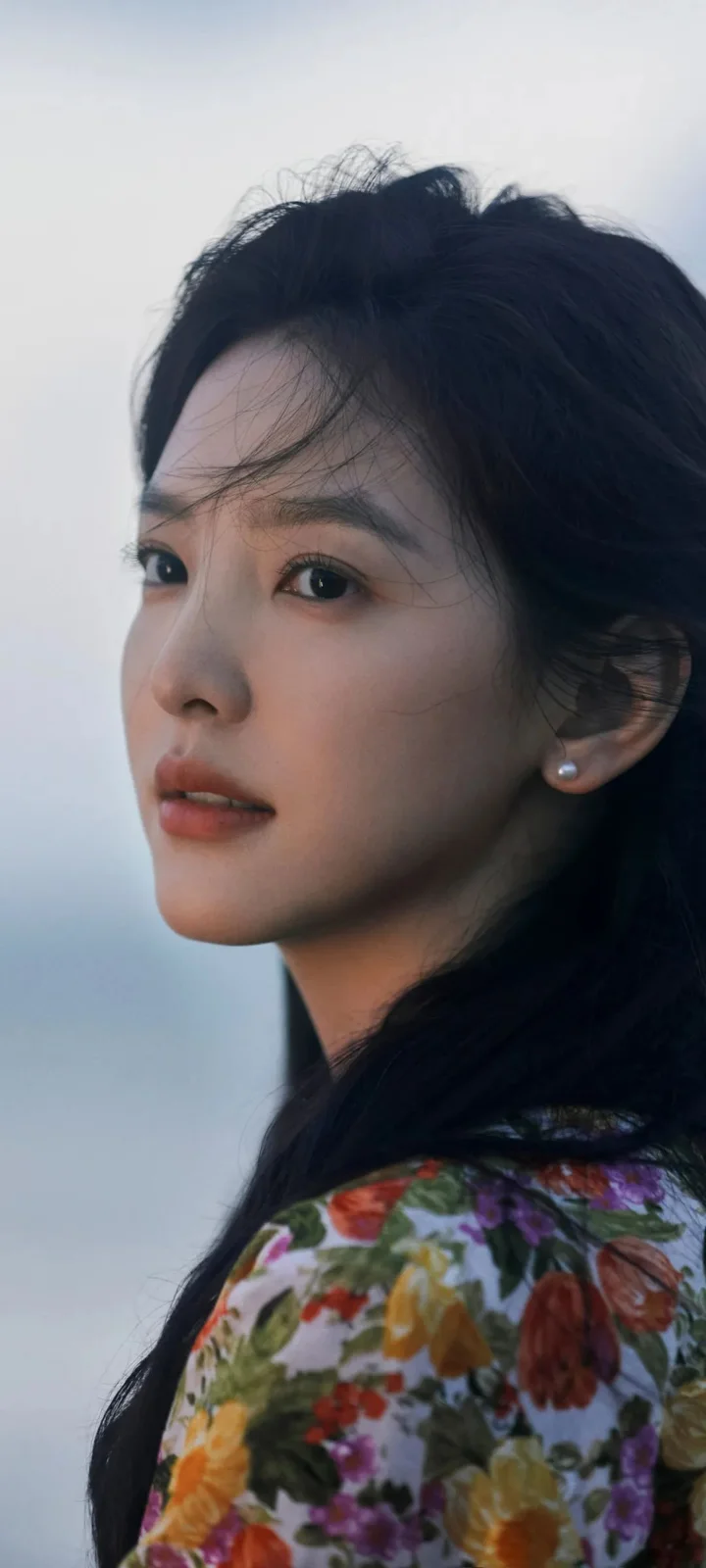 一位黑发女子身穿花卉图案上衣，面向侧前方，背景是浅色天空与海面。