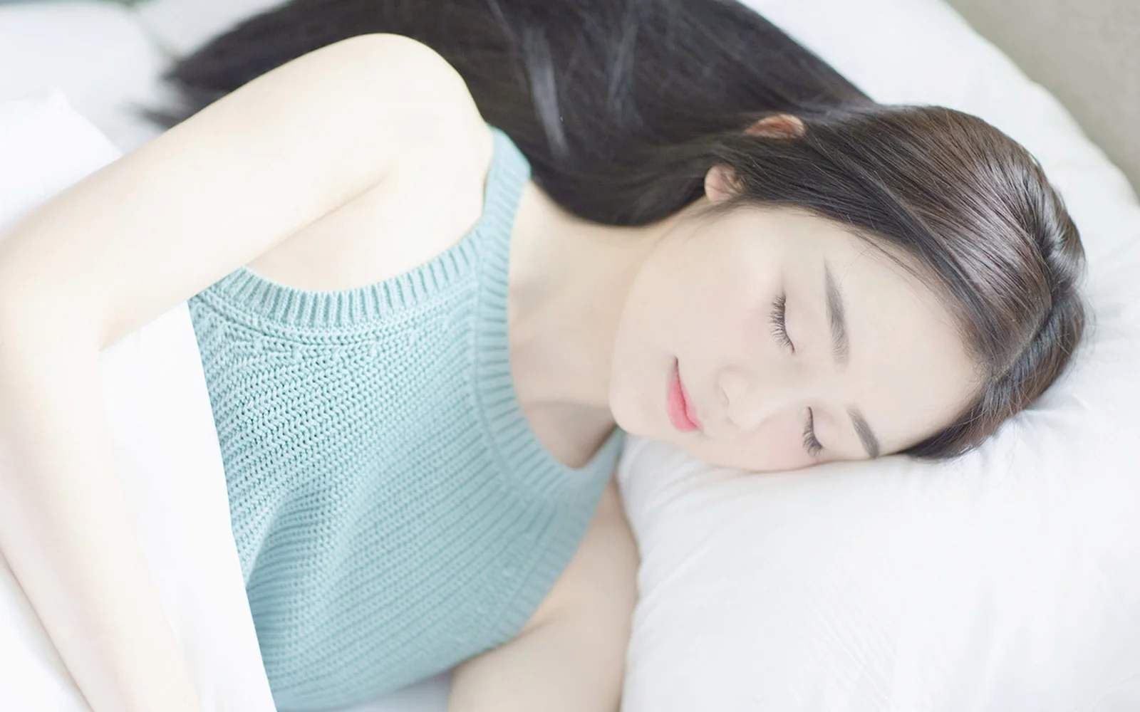 一名成年女性穿着浅蓝色针织背心，闭眼侧躺在白色床上睡觉。