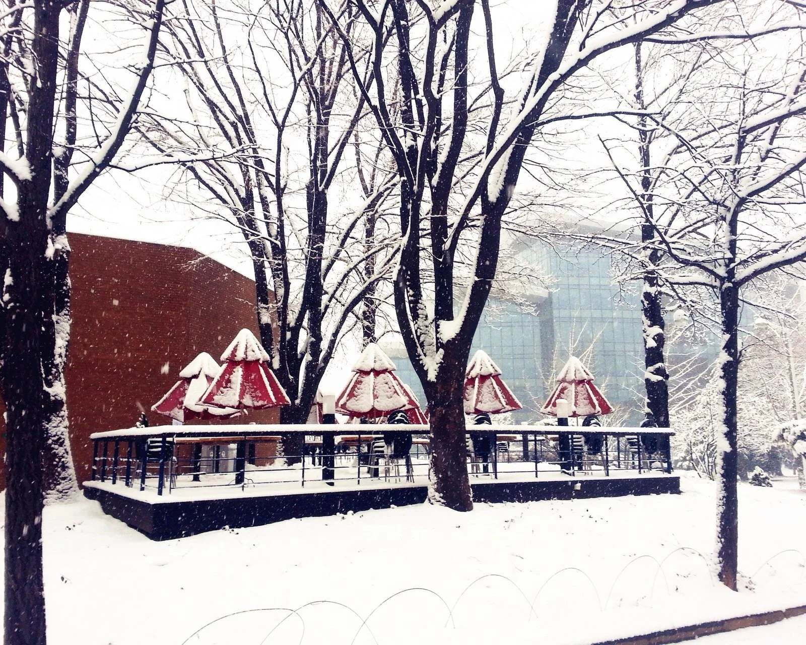 雪に覆われた公園で、赤い東屋が木々の間に並び、遠くにガラス張りの建物が見えます。