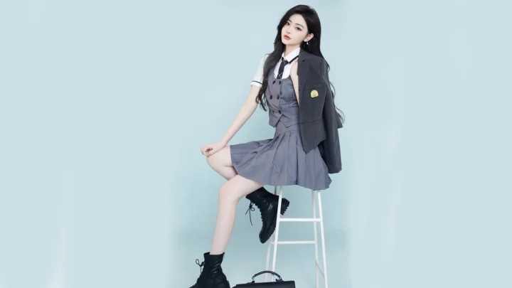 一名身穿灰色制服风格服装的女性坐在白色高脚凳上，背景为浅蓝色纯色墙面。