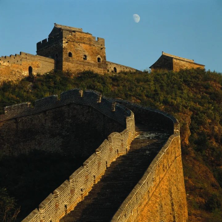 夕阳照亮的长城城墙与古堡建筑延伸在山脊上，蓝色天空中悬着一弯月亮。