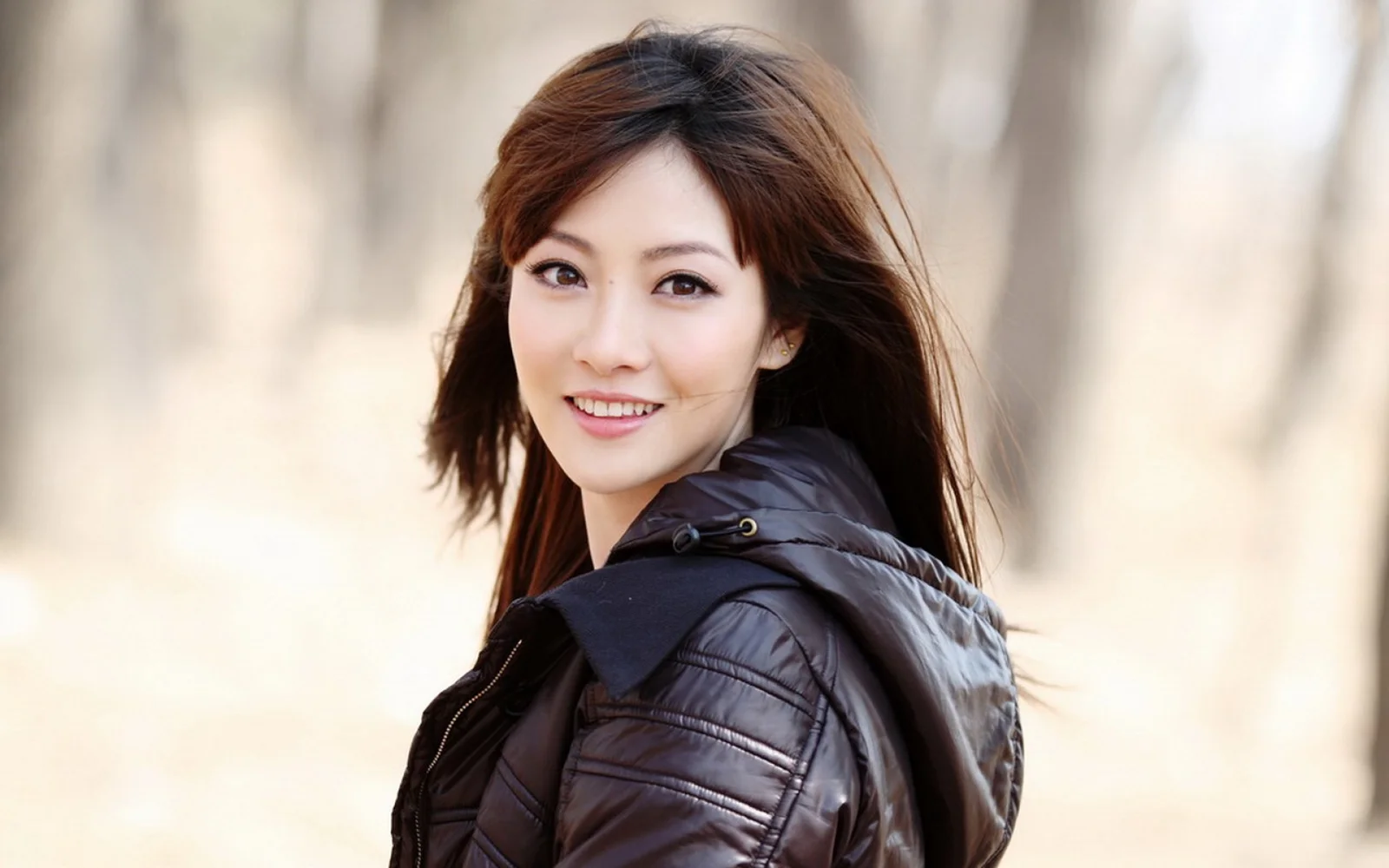 一位棕色长发女性穿着黑色夹克在户外微笑面对镜头。