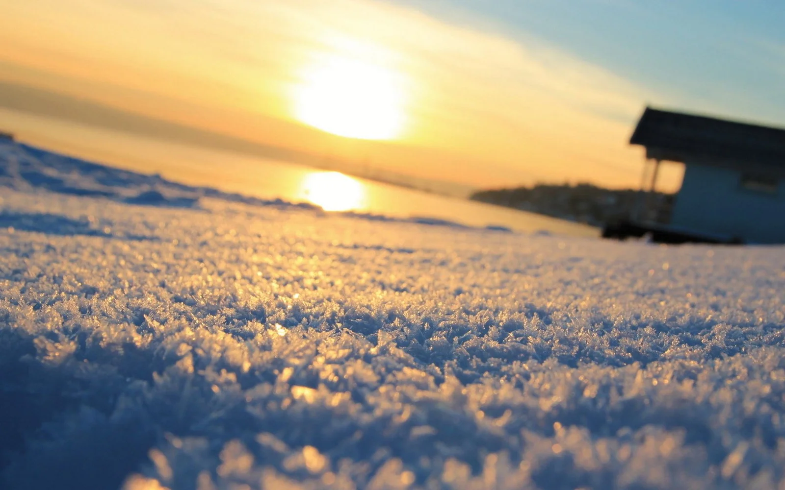 夕日が雪に覆われた湖畔を照らし、遠くに水面と小さな小屋が見えます。