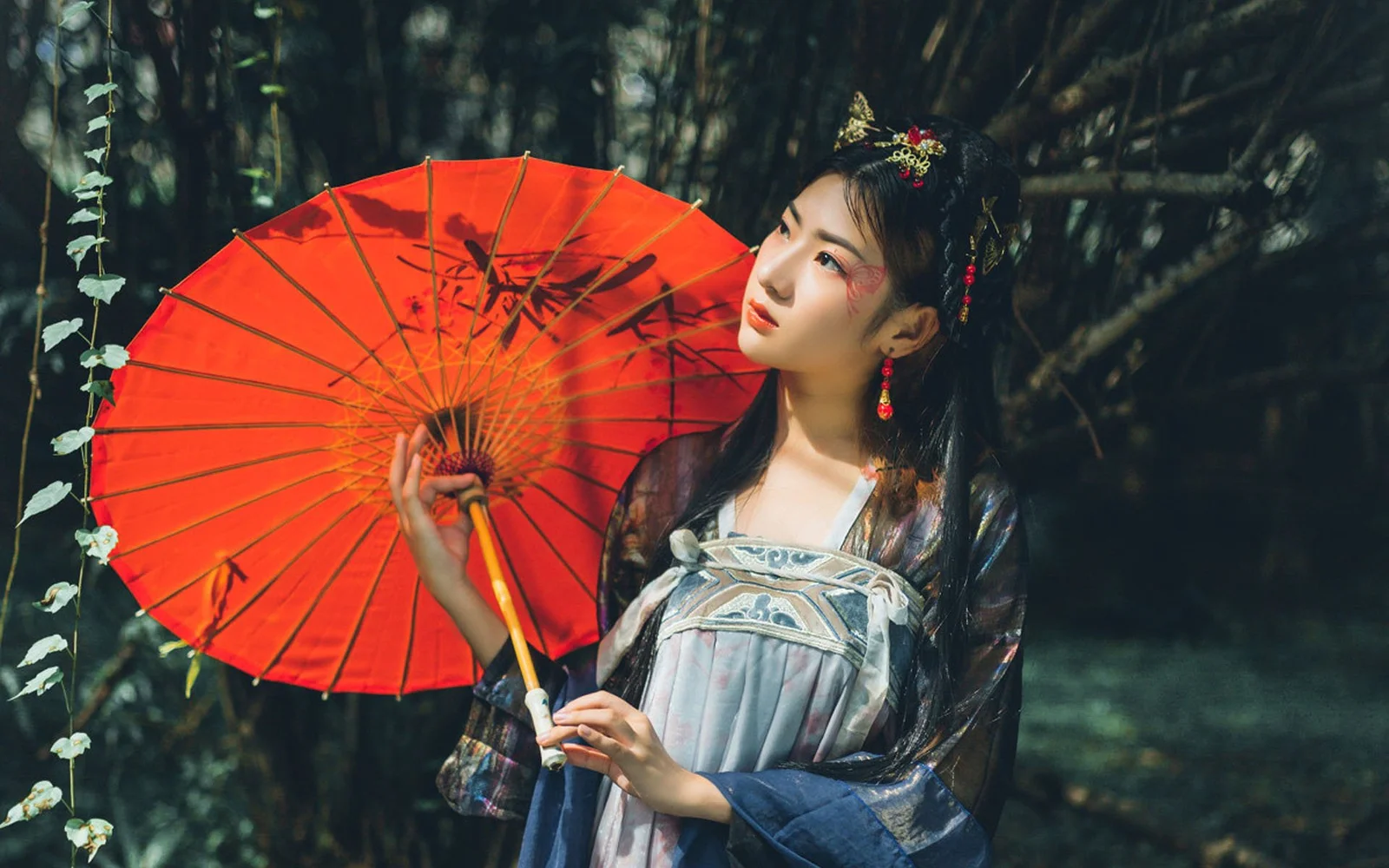 伝統衣装の女性が木々の中で赤い和傘を持っている。