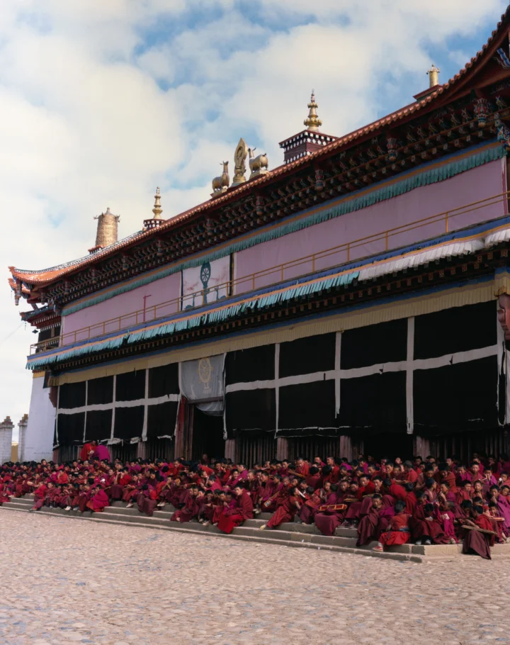 彩色藏式寺院建筑前坐着一群身穿深红色长袍的僧侣，天空中飘着云朵。