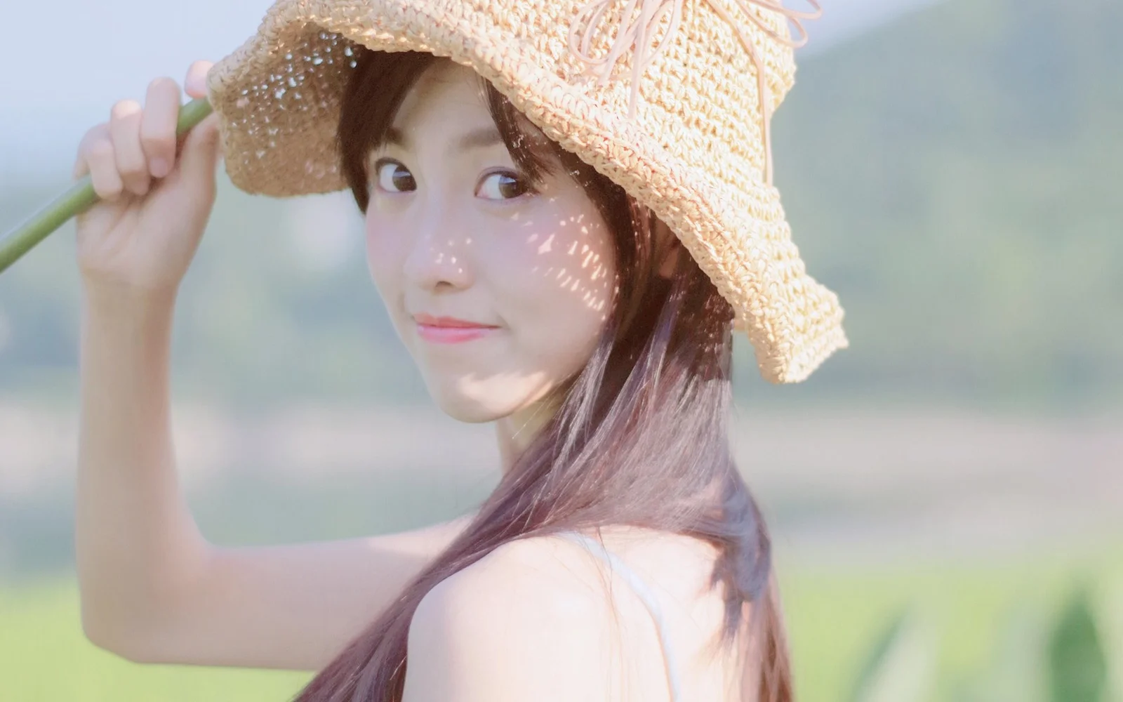 一名戴草帽的女性在户外回望镜头，背景是柔和的绿色景色。