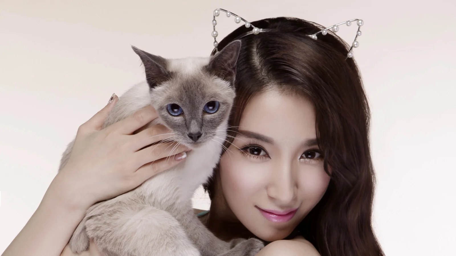 一名女子戴着猫耳发饰并抱着一只灰白色猫咪，背景为浅色。