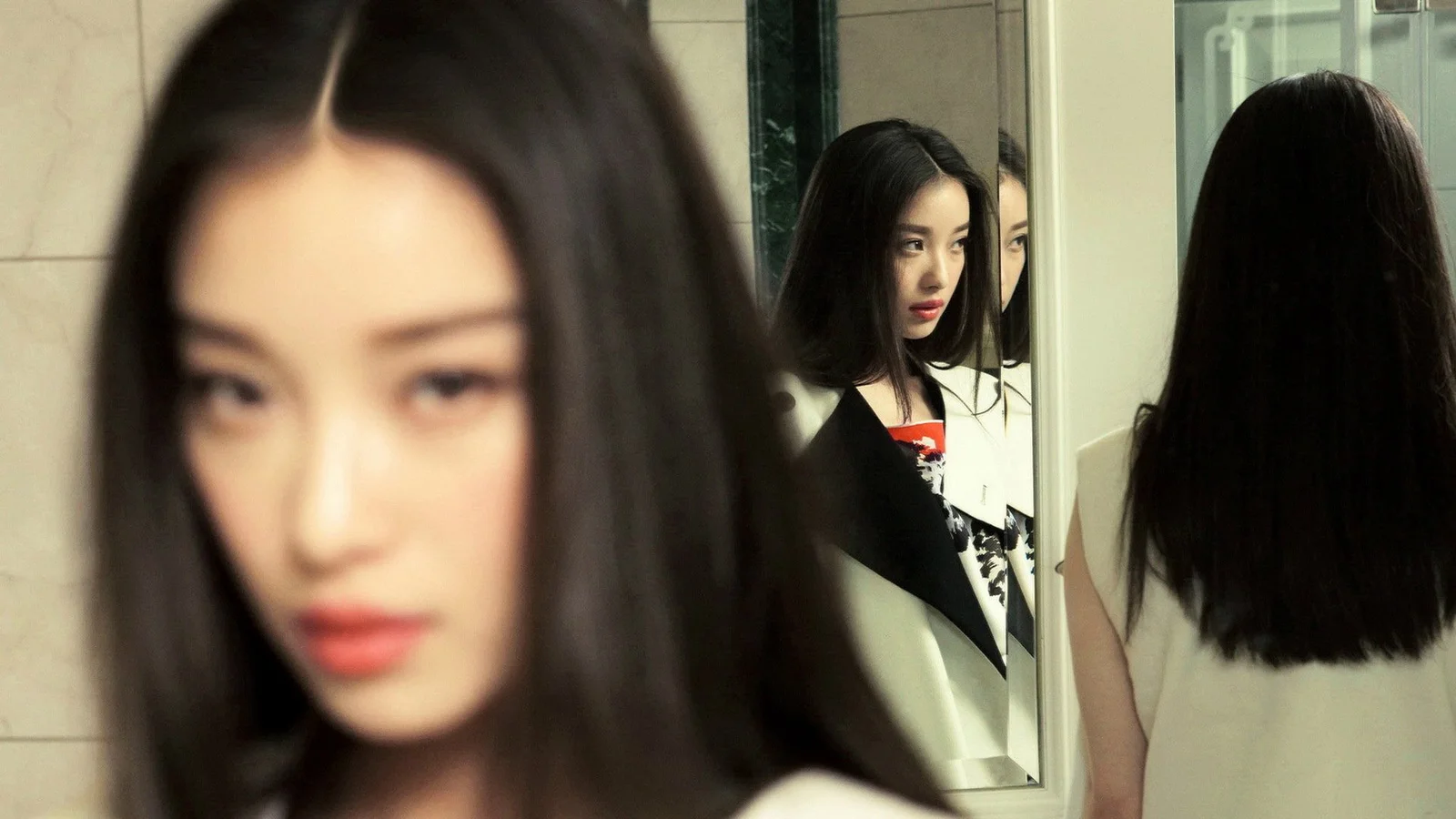 一名黑发女子站在镜子前，画面中呈现出她的模糊近景和镜中倒影。