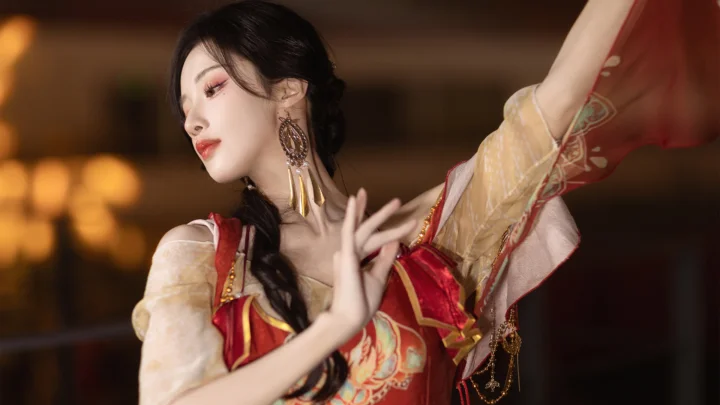붉은 전통 의상을 입은 성인 여성이 따뜻한 실내 배경 앞에서 춤 동작을 취하고 있다.