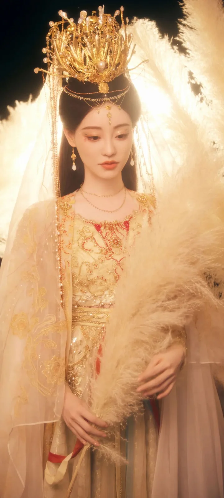 一名成年女子身穿金色华丽传统服饰，佩戴精致头饰，站在柔和的浅色羽毛背景前。
