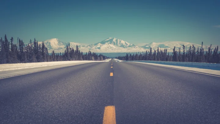 一条笔直的公路穿过积雪覆盖的森林，远处是壮丽的雪山。