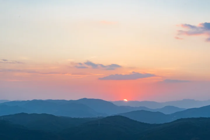 夕阳在层叠群山之间落下，天空呈现柔和的粉橙色渐变。