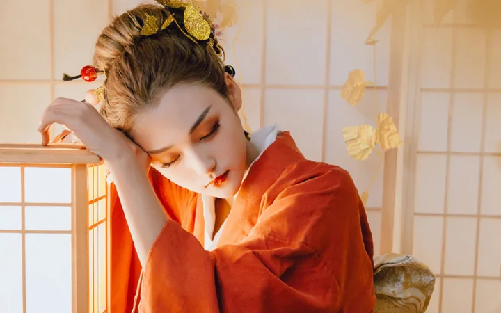 一名身穿红色传统服饰的女子在暖光窗边以手托头。