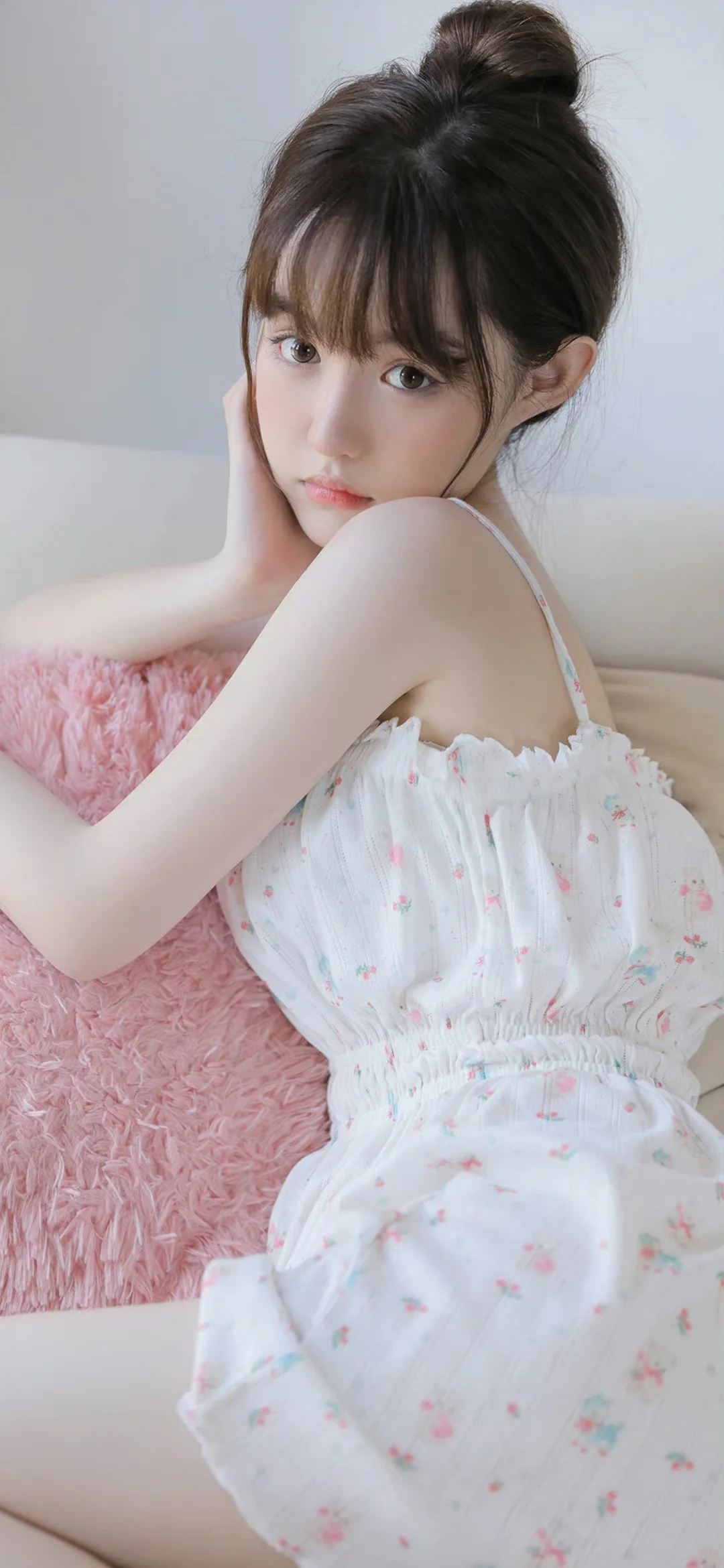 お団子ヘアの女性が白い花柄ワンピースを着て、ピンクの質感のある背景の前で頬に手を添えています。